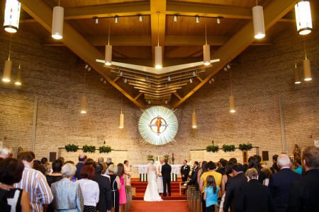 An image of a bride and groom standing at the altar in the center bottom portion of the photo before the priest at St. Basil the Great while the guests stand watching on the orange carpet and the middle to upper half of the photo shows the creamed colored brick walls with an unusual piece of artwork shaped in a white sunburst pattern with a painted image of Jesus standing in front of a cross in the middle of it and large wooden beams fill the top portion of the photo.