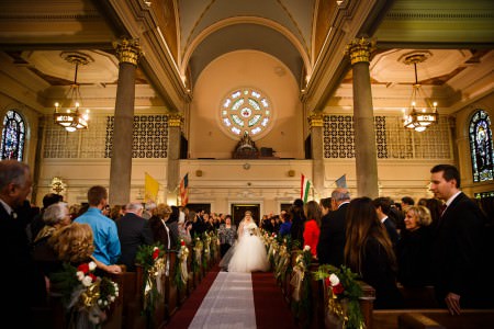 An image of a bride walking down the center aisle from the back of the church holding hands with her mom as they walk over a white aisle runner overtop of dark red carpeting, with the cream colored back wall of the sanctuary behind them with a large round stained glass window at the top center of the photo and the guests on either side standing and watching from the pews decorated with red and white roses bunched with greenery and gold bows at St. Elizabeth of Hungary.
