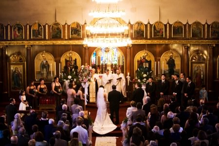 A bird's eye view of a bride and groom standing at the altar of the ornate St. Haralambos church where the carpet is deep red and the walls are ordained with holy painted images surrounded in ornate gold frames and the wedding guests are seated under a bright chandelier.