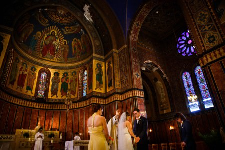 An image of a bride and groom facing each other holding hands in the lower right side of the photo with a priest and his raised hand praying over them inside the ornate sanctuary of St. James church decorated in dark browns, red, greens, and blues with some gold accents with paintings of saints on the arched and rounded walls and the left and purple colored stained glass windows on right.