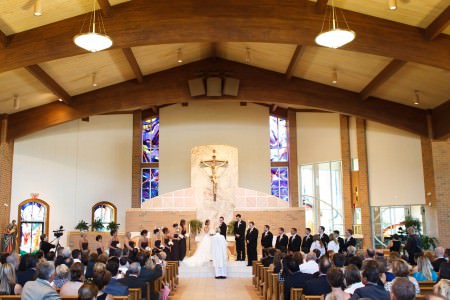 A photo taken inside the bright sanctuary of St. John Vianney church facing down the center aisle where the priest is standing at the altar with his back to the camera and a bride and groom are behind him facing him and the wedding party is lined up on either side the couple while the guests sit in the pews on either side watching and the background has a back cream colored wall with an arched wooden pillar lining the top of the photo and a large ceramic crucifix is attached to the wall with purple stained glass windows on either side.
