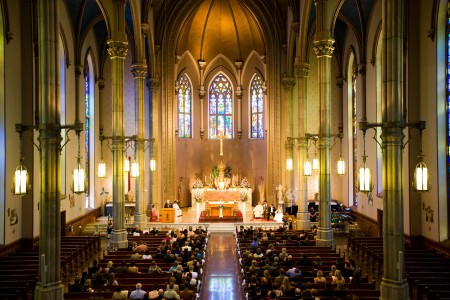 A photo taken inside the enormous sanctuary of St. Mary Catholic Church of a bride and groom sitting on the altar along with their witnesses and the priest while guests sit in the pews with large green pillars on either side of the photo and extremely tall cream colored walls with ornately arched stained glass windows in the upper half of the walls behind the altar.