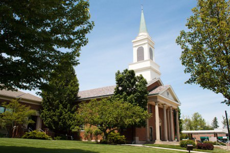 A picture of the outside of St. Mary of the Falls church from a side corner of the green yard where the main part of the brick church building white white pillars and a white steeple is centered in the right of the photo under a blue sky day with green trees up against the building as well as in the foreground on both upper sides of the photo.