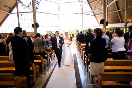A bride and groom walking arm and arm back down the center aisle in the center of the photo with a wall of windows full of bright white light behind them and the wedding guests standing at the wooden pews on either side of the aisle at St. Paschal Baylon Parish.