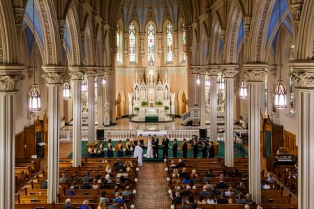 An image taken from the balcony of St. Patrick Parish overlooking a bride and groom standing arm and arm facing the congregation on a dark green carpeted platform with the wedding party standing on either side of the them in the ornate sanctuary full of tall arched light colored beams and blue painted ceiling inside the arched beams and tall thin stained glass windows above the altar and guests seated in the wooden pews facing the wedding party.