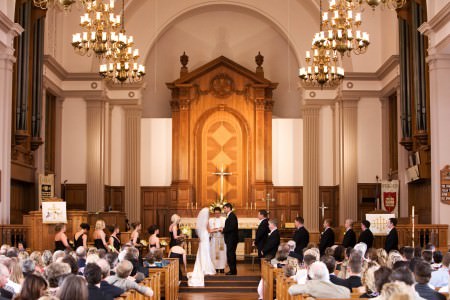 A picture taken from the back of the sanctuary of a bride and groom standing at the altar during the wedding ceremony at St. Paul's Episcopal church with tall pillars, an ornate wooden altar wall and high ceilings.