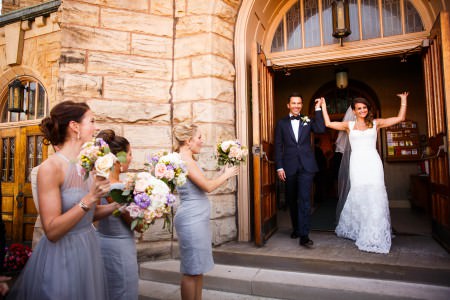 A bride and groom holding hands up high with smiles exiting St. Vincent church with double wooden door and stone walls with the bridesmaids in grey blue dresses holding purple and pink bouquets cheering them on.