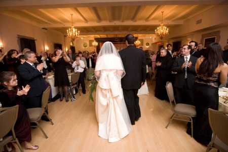 An image from behind of a bride and groom entering the reception hall walking hand in hand in the center of the wedding guests standing at the tables clapping at the Metropolitan Centre.