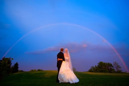 A stunning shot of behind a bride and groom standing side by side with their arms around each other waists on a grassy hill looking out at a rainbow arching right over them on an extremely blue sky at dusk.