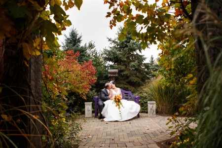 A bride holding a bright fall colored bouquet leaning into her groom for a kiss while they are sitting on a purple bench set on a round brick patio in the middle of a garden with fall foliage at the Thorncreek Winery.