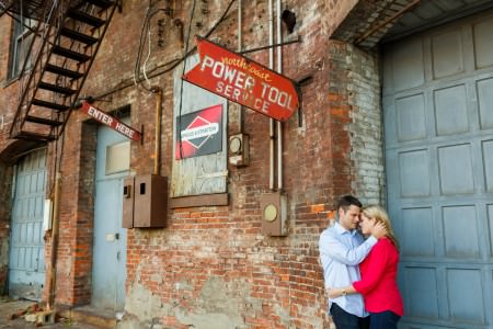 An image taken outside up against an industrial looking brick building of an engaged couple with their arms around each other and leaning with their foreheads pressed against each other in the lower right corner of the photo where the corner of a brick building meets a blue garage door and the upper left of the photo has red signs and a wrought iron escape stairs attached to the building.