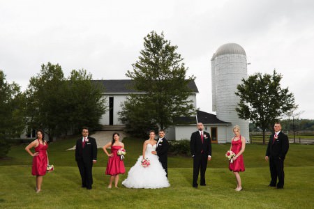 A bride holding her red and white bouquet as her groom holds her waist standing on a grassy knoll with their wedding party in black tuxedos and red dresses standing with them and a white barn and silo in the background on an overcast day at Walden Inn.