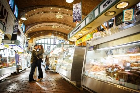 An engaged couple with jeans and olive green colored tops stand embracing with their heads together on a brick floor inside a bakery row of the Westside Market in Cleveland with an brick arched wall in the background with a wall of windows and bakery and food display cases on either side of the couple.