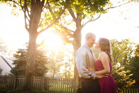 An image of an engaged couple standing nose to nose with their hands on each other's waists where the woman has on a fuchsia strapless dress and the man is wear a grey button down shirt with matching tie while standing outside in a residential backyard with a brown picket fence in the background with green trees and shrubs while the bright sun shines down on the couple creating a beautiful sunny haze.