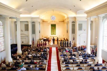 A balcony view of a bride and groom holding hands at the altar with their wedding party standing looking on inside the white pillared sanctuary of the Westminster Presbyterian church with red carpeting and a white aisle runner.