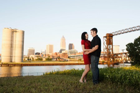 An engaged couple stand on a patch of grass in the right of the photo looking into each other's eyes with their hands on each other's waists where the woman is wearing a short red dress and the man is wearing a black golf shirt and jeans with the river and the Cleveland skyline is in the background on a sunny evening.