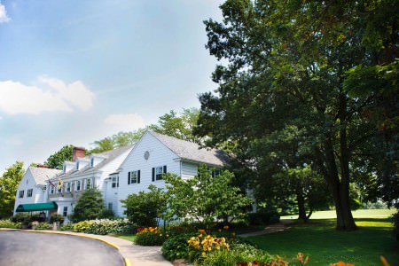 A picture of the outside of the Wooster Inn taken from the curved driveway on a sunny summer day where the building is in the center left of the photo with green grass and yellow and red flower beds and tall green trees are on the right.