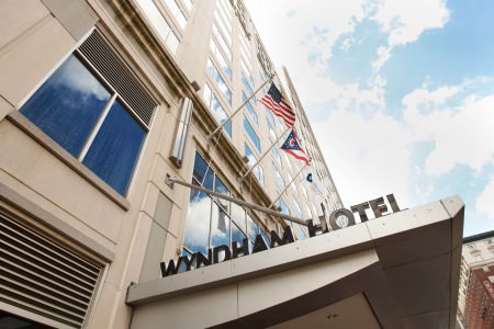 A picture taken from the sidewalk looking up the sand colored stone block building of the Wyndham Hotel with the words of the hotel name are in black block letters on the top edge of the entrance overhang with flags hanging from the building above the letters on a bright blue clouded with the sky reflected in the rectangular windows of the building.