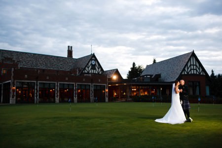 A picture taken of a bride in her long white gown with a long veil standing with her groom dressed in kilt in the far right of the photo on the green front lawn of the Tudor and red brick style building of the Youngstown Country Club taken at dusk with a golden light shining from the building all under a grey cloudy sky.