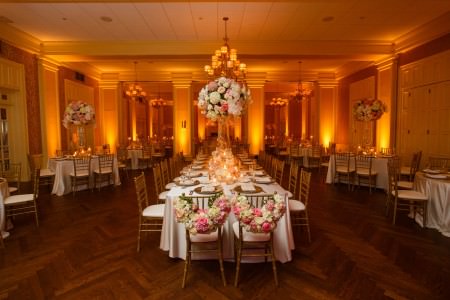 An image of a reception room at Brookside Country Club in Ohio is decorated with golden uplights where rectangular tables surround the outer walls and a large head table is set in the center of the room all with gold and white chairs and the bride and grooms chairs are placed at the head of the table decorated with arched pink and white flowers.