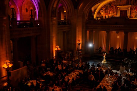 A bird's eye view of a bride and groom dancing at their reception inside the darkened ballroom of the Old Courthouse set with golden orange up lights, marble pillars, and vaulted ceilings and the guests are all seated.