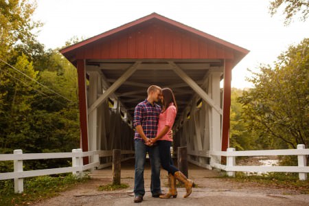 An engaged couple stand holding hands and resting forehead to forehead on the path with a red covered bridge surrounded by white fencing.