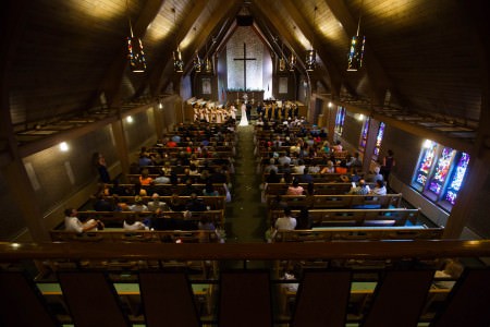 A bird's eye view of a wedding ceremony taken from the balcony of Greensburg United Methodist Church looking down the center aisle carpeted in olive carpet with white flower petals dropped and wooden pews on either side filled with guests watching the bride and groom standing with the pastor and the wedding party in front of the altar with a white stone wall and a large wooden cross all inside a sanctuary of wooden and brick walls with stained glass windows on the right.