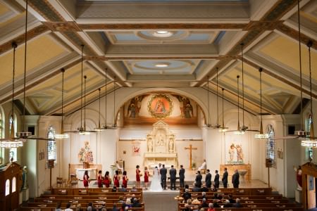 A photo taken from the balcony of the Holy Rosary Church overlooking a bride and groom facing the altar with the bridesmaids on the right in red dresses and the groomsmen on the left in black tuxedos in the bright sanctuary with the walls and square ceiling patterns in yellow and blue.