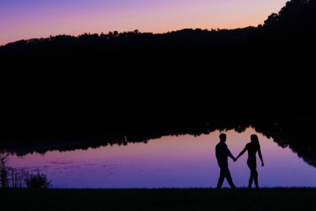 A photo of an engaged couple's black silhouette walking in front of Indigo Lake in Ohio at dusk where the lake is reflecting the purple and pink hued night sky.