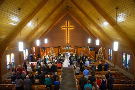 A photo of a wedding ceremony where the bride and groom are standing in the center of the photo at the end of the center aisle facing each other and holding hands while the guests stand up from the wooden pews under a tall triangular ceiling made of wooden planks and curved wooden beams with a brick wall behind the altar with a large wooden cross lit with yellow lights from behind.