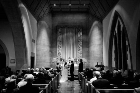 A black and white image of a bride and groom standing facing each other at the altar of St. Paul's Episcopal Church with the priest in the center facing the camera and the maid of honor to the left of the bride and the best man to the left of the groom with tall candelabras light on the altar with a tall two story stone wall behind them and guests seated in the wooden pews on either side of the aisle.