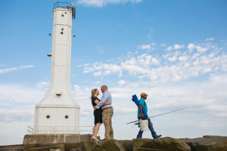 An image of an engaged couple where the woman is in a black dress with white high heels facing the man while they are hugging and he is wearing tan pants and a blue button down shirt standing at the bottom of the photo on large boulders with an old white light house to the left of the couple and a fisherman walking away from the couple on the right carrying a white bucket, a fishing pole, and a blue camping chair slung over his arm all under a blue sky with white puffy clouds in Huron, Ohio.
