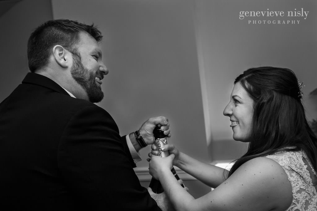 Bride and groom share a look while popping the cork out of a bottle of champaign. 