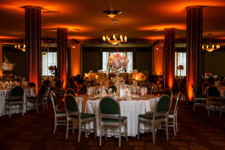 An image of a room at The Silver Grille decorated for a wedding reception where a round table with green and silver chairs are at the focal point in the center of the photo white tablecloth and pink linens and a pink and white floral arrangement is set on a tall silver holder and the background of the photo shows four pillars light up with orange glowing lights that shine up towards the ceiling.