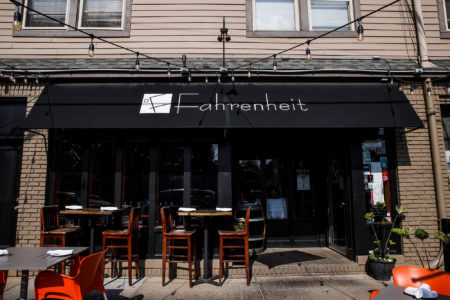 An image of the outside of the Fahrenheit restaurant with the brown painted brick walls and black awning hanging over their outdoor seating area with tall wooden chairs and white napkins set at the tables.