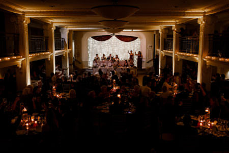 A picture of a wedding reception set in a the darkened ballroom at Park Lane where the guests are seated at round tables on the ballroom floor raising their glasses in a toast while the wedding party is at tiered rectangular tables with white twinkle lights under tulling behind them.