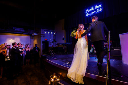 An image taken in a darkened room at the Music Box Supper Club where a bride and groom are stepping up together onto the stage in the right side of the photo under purple and blue lights and guests are standing and clapping for them on the right side of the photo.