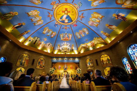 A bride and groom stand facing the altar inside the ornate Holy Trinity Greek Orthodox Church in Ohio with a round domed ceiling painted with a blue background and angelic images and stained glass windows.