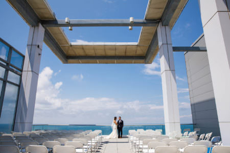An image of a bride in her white dress standing with her groom against the glass railing on a sunny summer day with blue skies and puffy clouds and a view of Lake Erie behind them from the balcony set with white folding chairs lined up for the ceremony at the Aloft building in downtown Cleveland, OH.