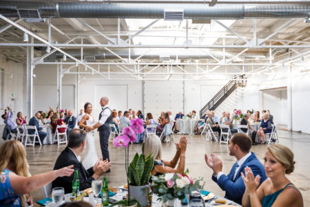 A wedding reception in a spacious, industrial-style venue known as The Madison with guests seated at round tables applauding the bride and groom dancing in the center. Bright, airy atmosphere with visible ductwork overhead