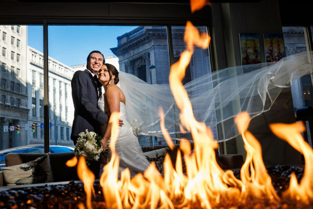 A bride and groom smile joyfully near a window with a cityscape backdrop, the bride's long veil dramatically caught in the breeze above a decorative fire pit at their Cleveland wedding.