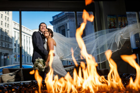 A bride and groom smiling, standing close together inside a room at the Kimpton Schofield Hotel with flames in the foreground and a city street view through the window behind them. The bride's veil is